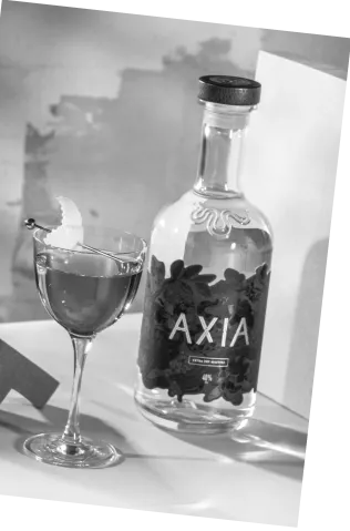 Black and white picture of an Axia Greek Mastiha Spirit x Gaucho Sweet Vermouth Narcissus Cocktail on a counter with an Axia Spirit bottle next to it and someone holding an olive