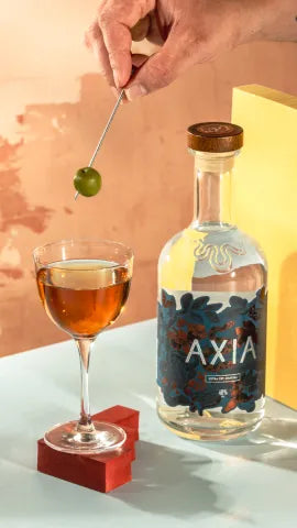 Axia Greek Mastiha Spirit x Gaucho Sweet Vermouth Narcissus Cocktail on a counter with an Axia Spirit bottle next to it and someone holding an olive