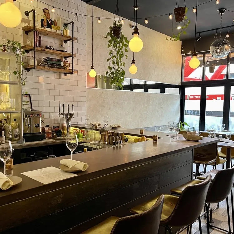 Pomp and Circumstance, New York: warmly lit, modern bar and dining area with a dark wooden bar, leather stools, pendant lights above and tables set with plates and glasses