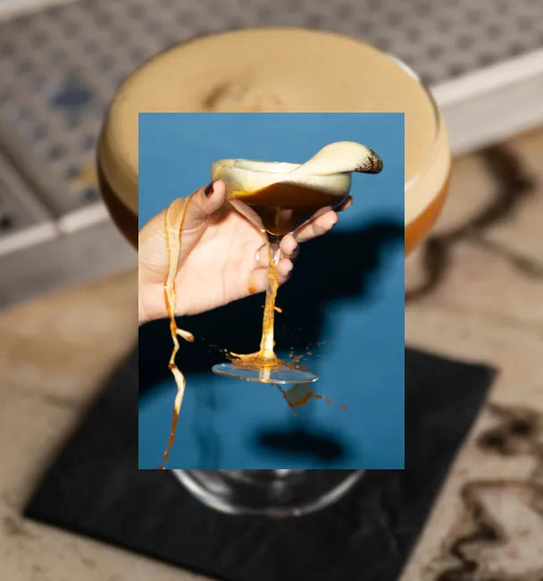 Hand holding Axia Spirit Axpresso Martini Cocktail, spilling out of glass, on blue background with secondary blurred picture in the background of another Axpresso Martini