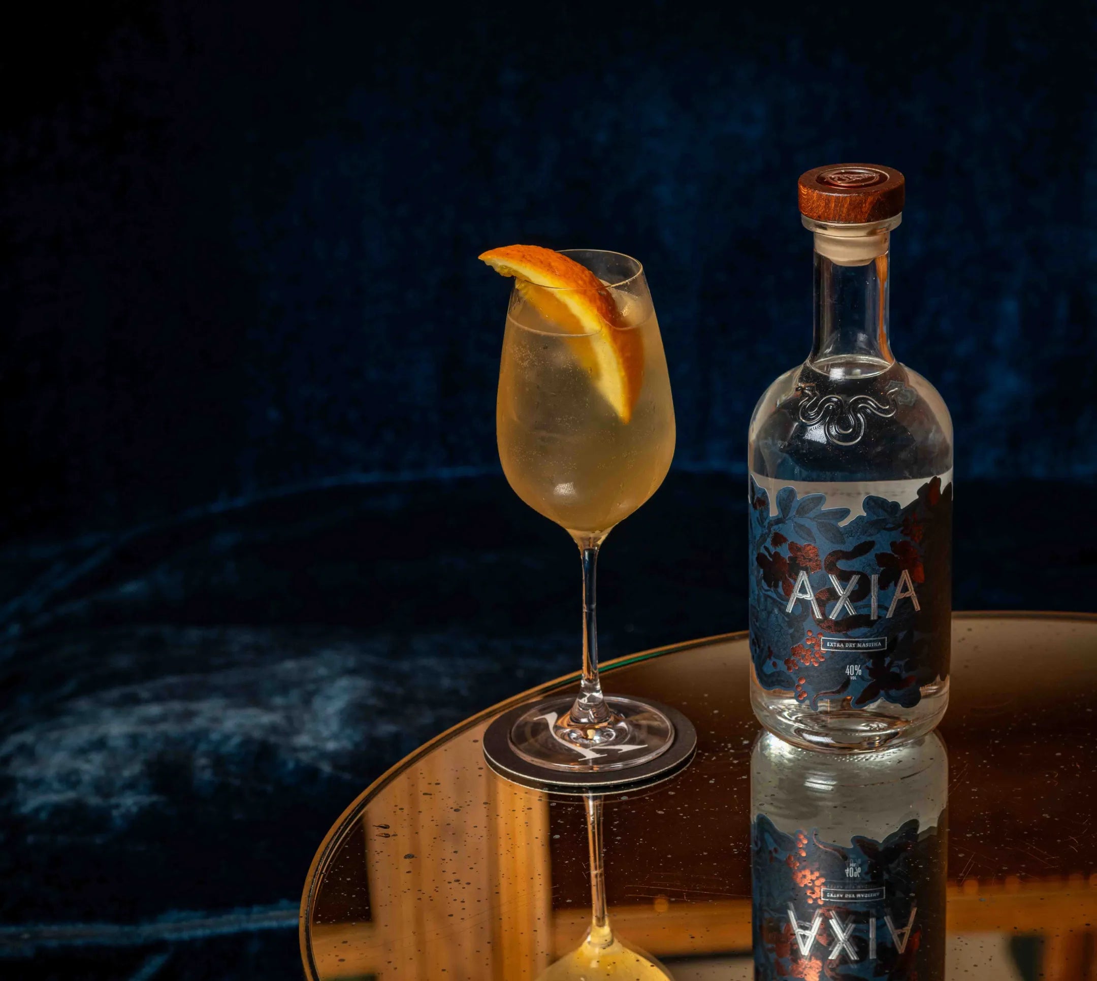 Bottle of Axia Mastiha Spirit next to a cocktail glass with orange slide on a mirror table