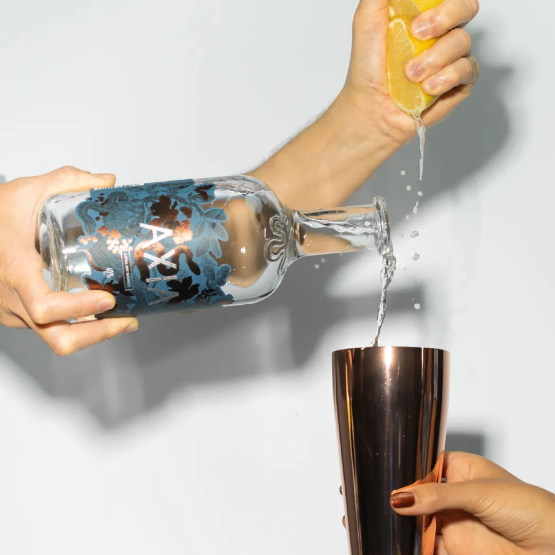 Hand holding a bottle of Axia Mastiha Spirit pouring it into a metal cocktail shaker and squeezing lemon juice with the other hand