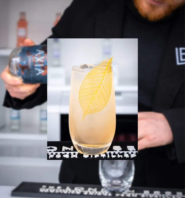 Axia Spirit and London Essence Soda Yamas Cocktail on a bar with a secondary blurred picture in the background of a bartender making an Axia cocktail