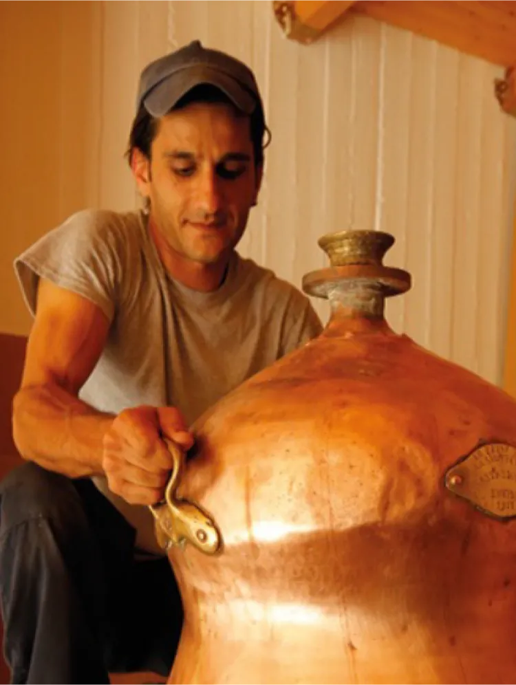 A person wearing a grey t-shirt and a cap, crouched down, holding the brass handle of a large copper still for mastiha spirit blending process