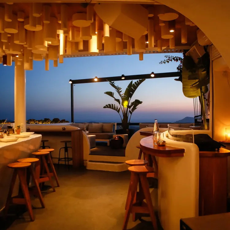 Tango Champagne and Cocktail Bar, Santorini: chic outdoor bar at dusk with white curved counter, wooden stools, lounge seating, sea view, and hanging lights