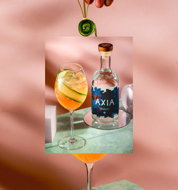 Axia and Gaucho Watermelon Spritz cocktail next to an Axia Mastiha Spirit bottle with a secondary blurred picture in the background of another Watermelon Spritz cocktail