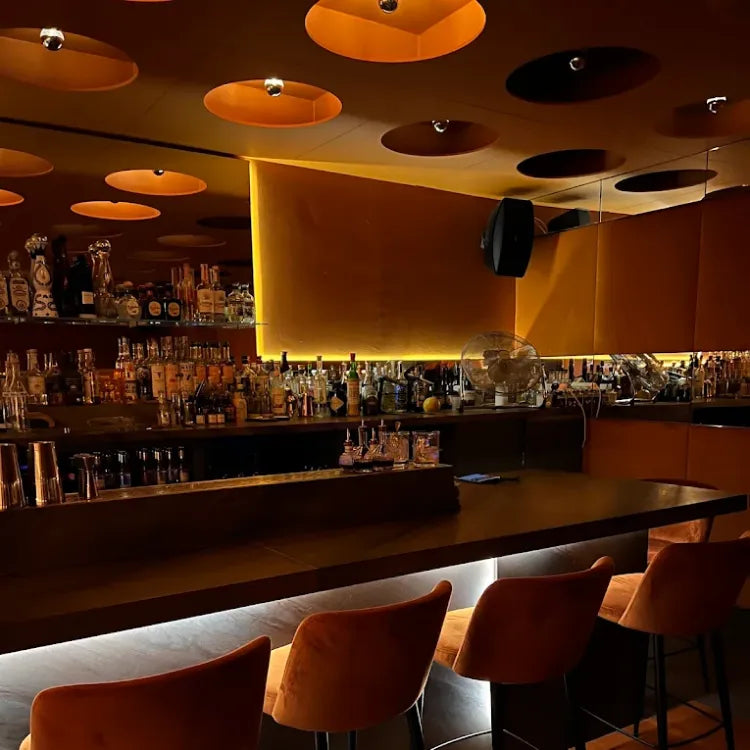 Zēphyr Notting Hill, London: warmly lit bar with long counter, several orange velvet bar stools, circular orange and dark brown light fixtures and well-stocked bar