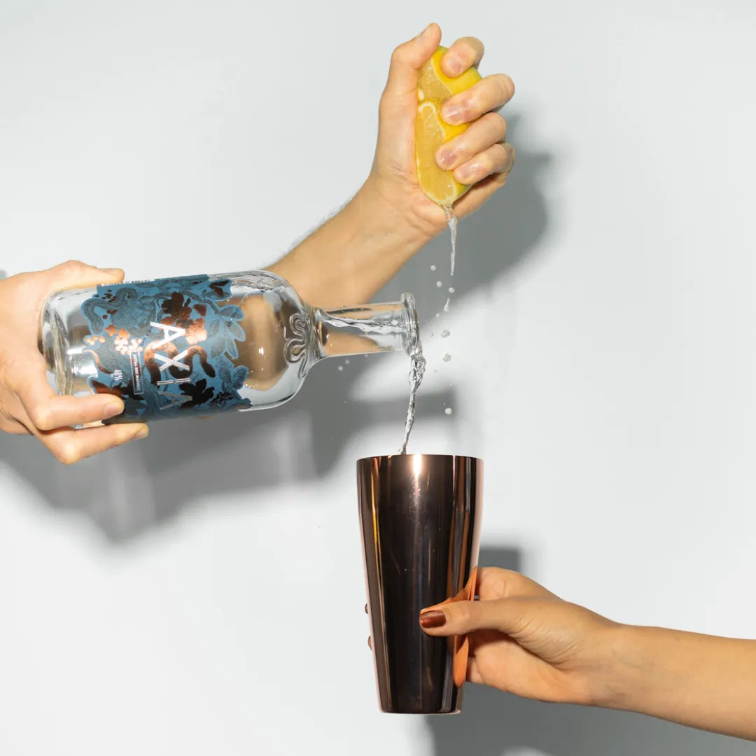 On a white background, hand pouring Axia Mastiha Spirit in a metallic cocktail glass while the other hand is pressing a lemon into it