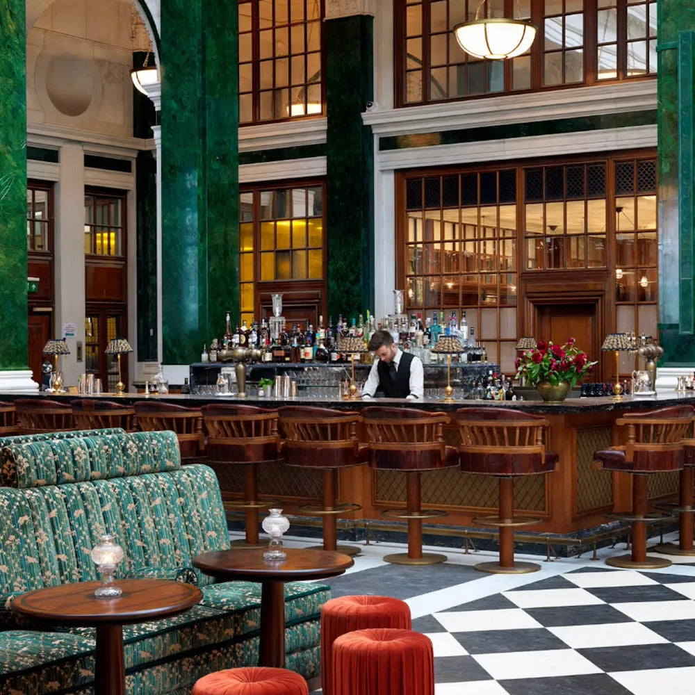 The Ned, London: Elegant restaurant bar with bartender, checkered floor, green columns, decorative tables, and plush seating.