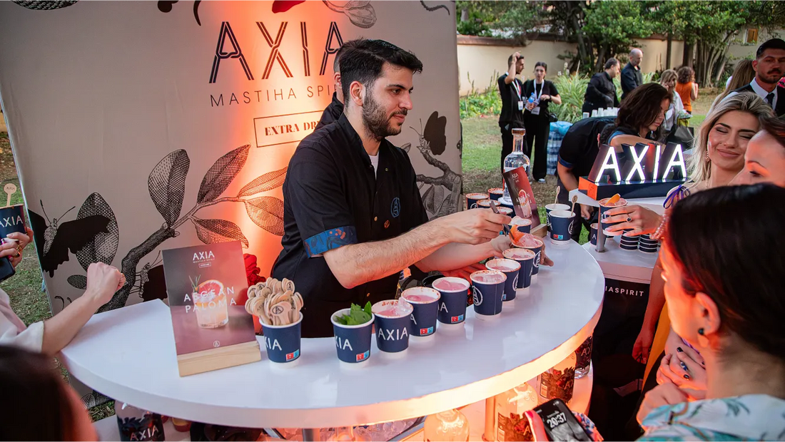 Bartender Babis Baldoumis serves Aegean Paloma cocktails at an Axia Mastiha Greek Spirit event booth, surrounded by guests and branded decorations