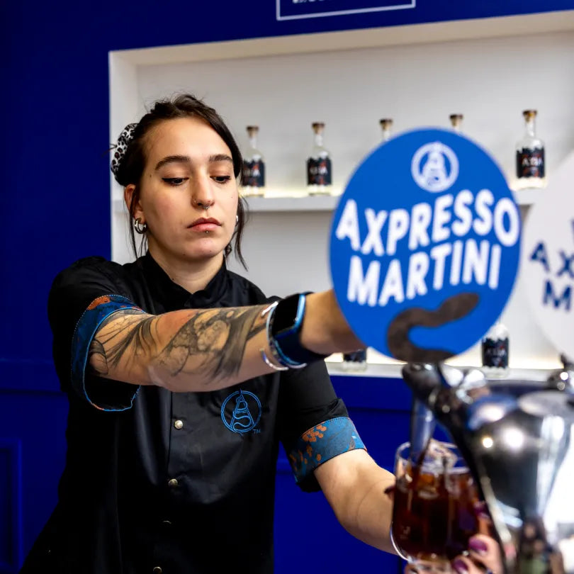 Bartender pouring an Axia Mastiha Spirit cocktail drink at a bar with 'Axpresso Martini' branding above it