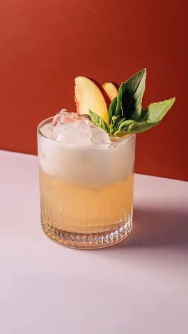 Axia Greek Mastiha Spirit Basilisk Smash Cocktail with ice, peach slices and basil on a red background
