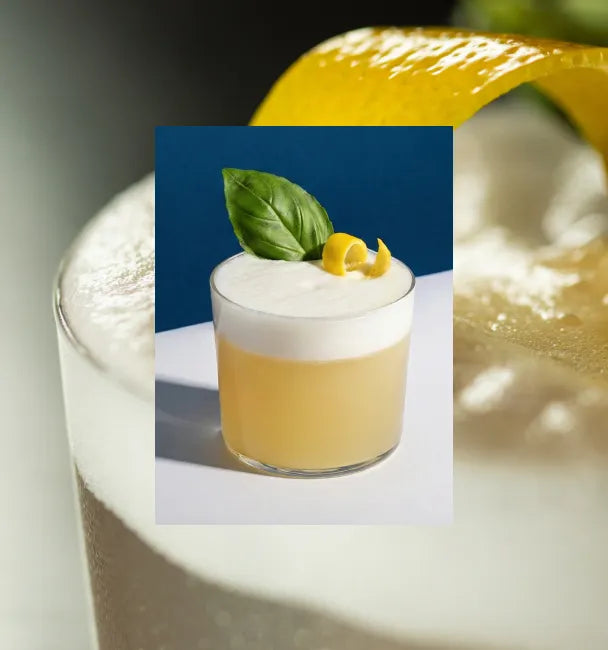 Axia Greek Mastiha Spirit Sour Cocktail with foam, basil leaf and lemon peel on a blue background with a secondary picture in the background of another Sour cocktail