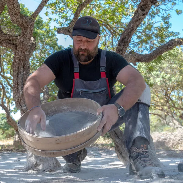 A bearded man in a black t-shirt and grey overalls kneels outdoors, sifting material through a large sieve amidst mastic trees