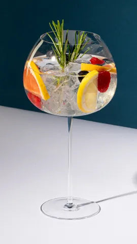 Axia Greek Mastiha Spirit Tonic Cocktail garnished with lemon slices, raspberries, and rosemary on a dark blue background