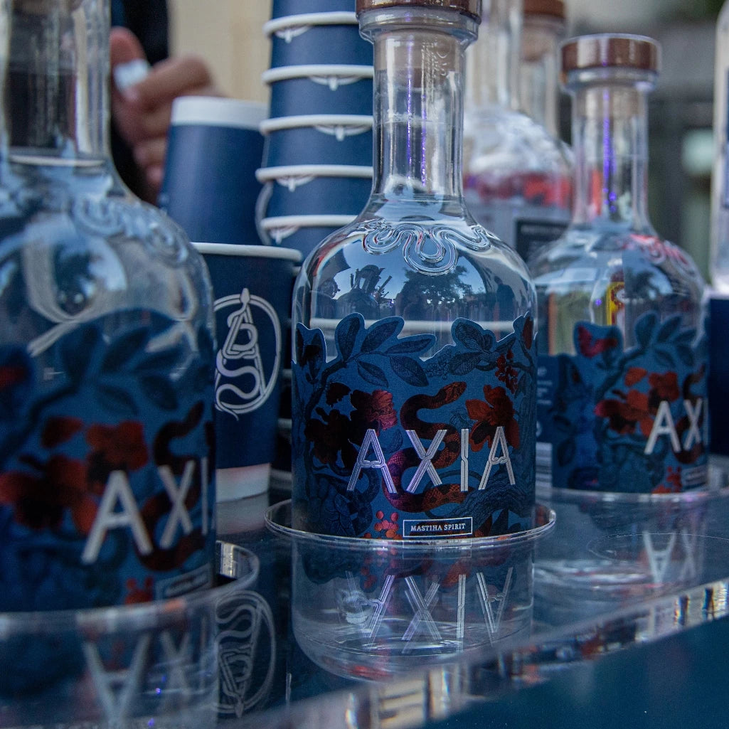 Bottles of Axia Greek Mastiha Spirit displayed on a reflective surface alongside in front of stacked dark blue Axia drinking cups