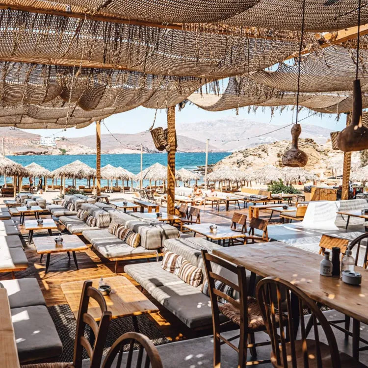 Alemagou, Mykonos: Outdoor seating area with tables, chairs, and couches by a beach.