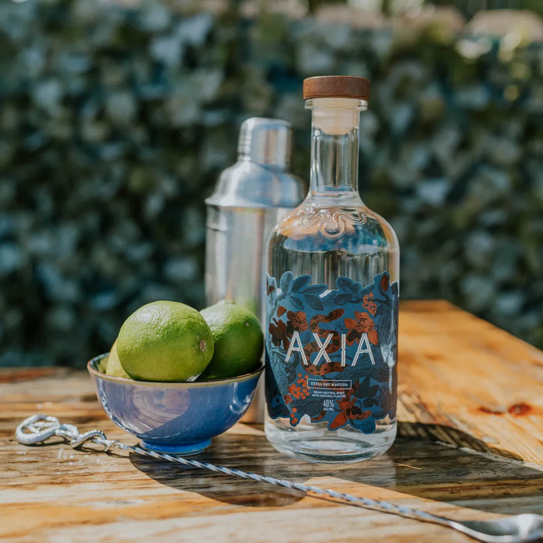 Bottle of Axia Mastiha Spirit on a wooden table outside next to a bowl of lemon and an Axia Cocktail Stirrer