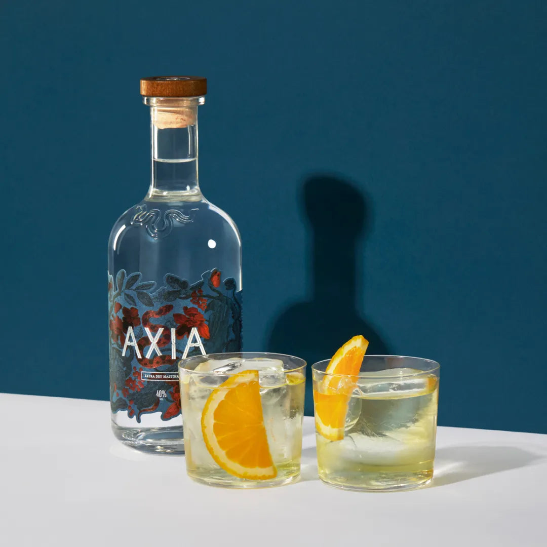 Bottle of Axia Spirit next to two cocktails with a slice of orange each on a white table and dark blue background